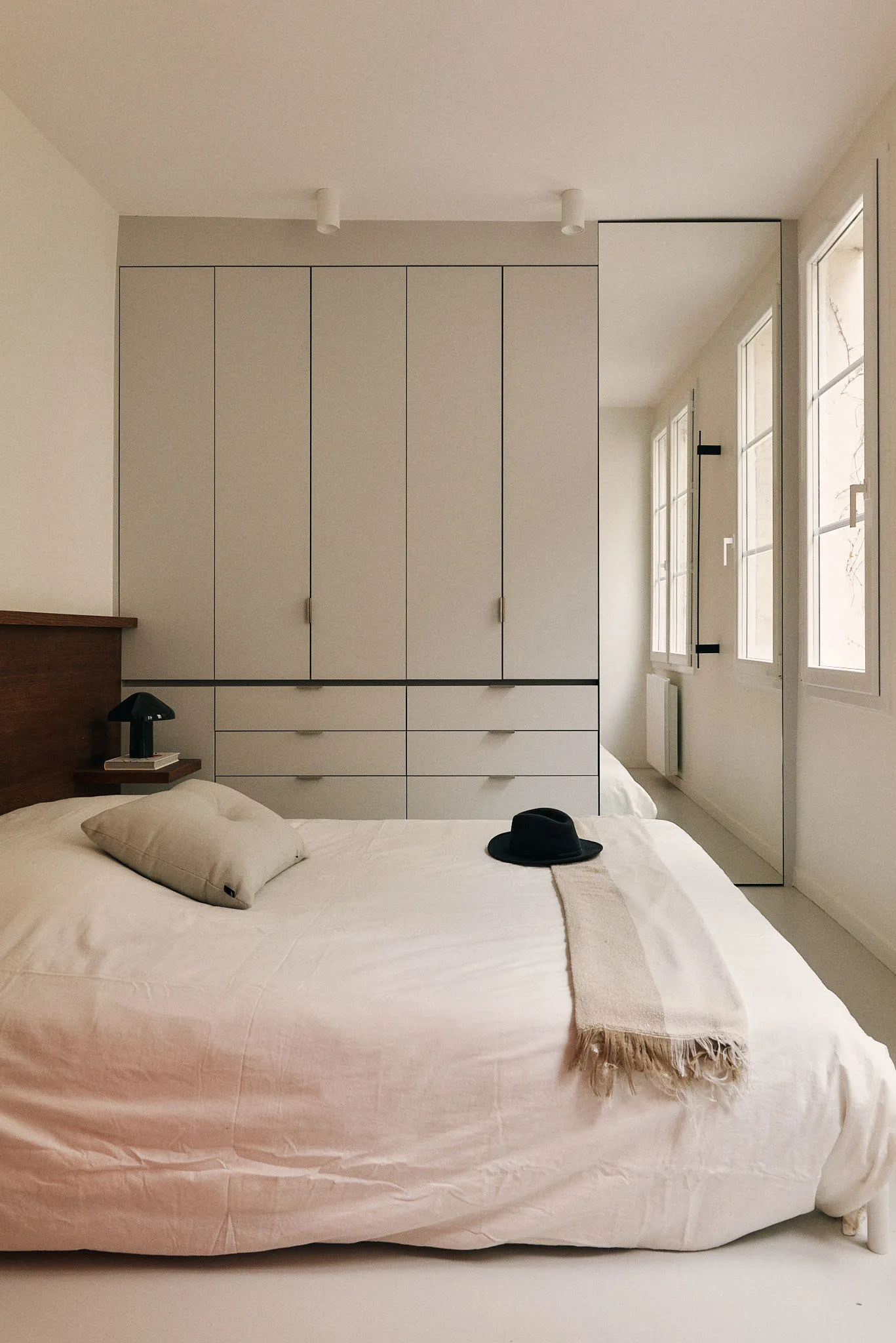 A modern minimalist bedroom with concrete floors, white bedding, and a large window allowing natural light to flood the space