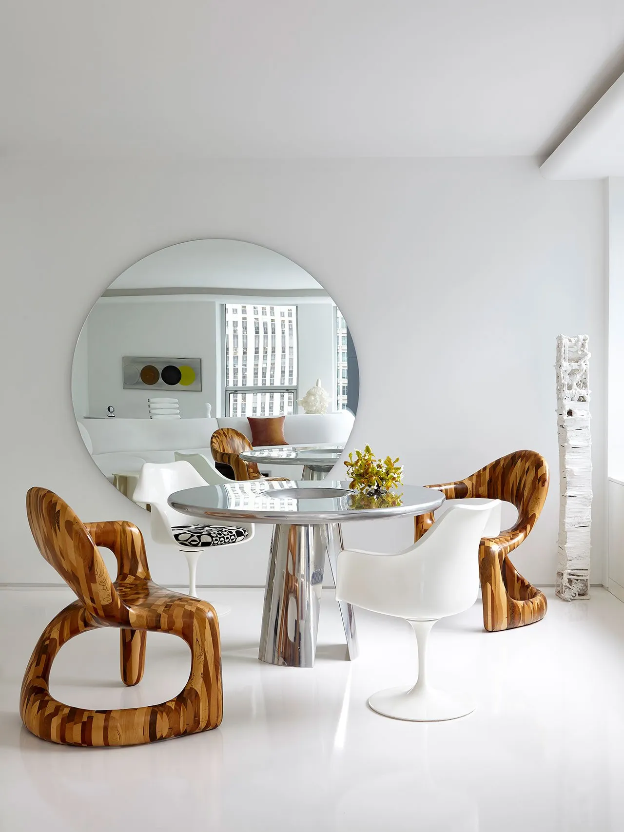 A modern contemporary living room featuring metal, wooden, and plastic furniture with white walls and a large circular mirror