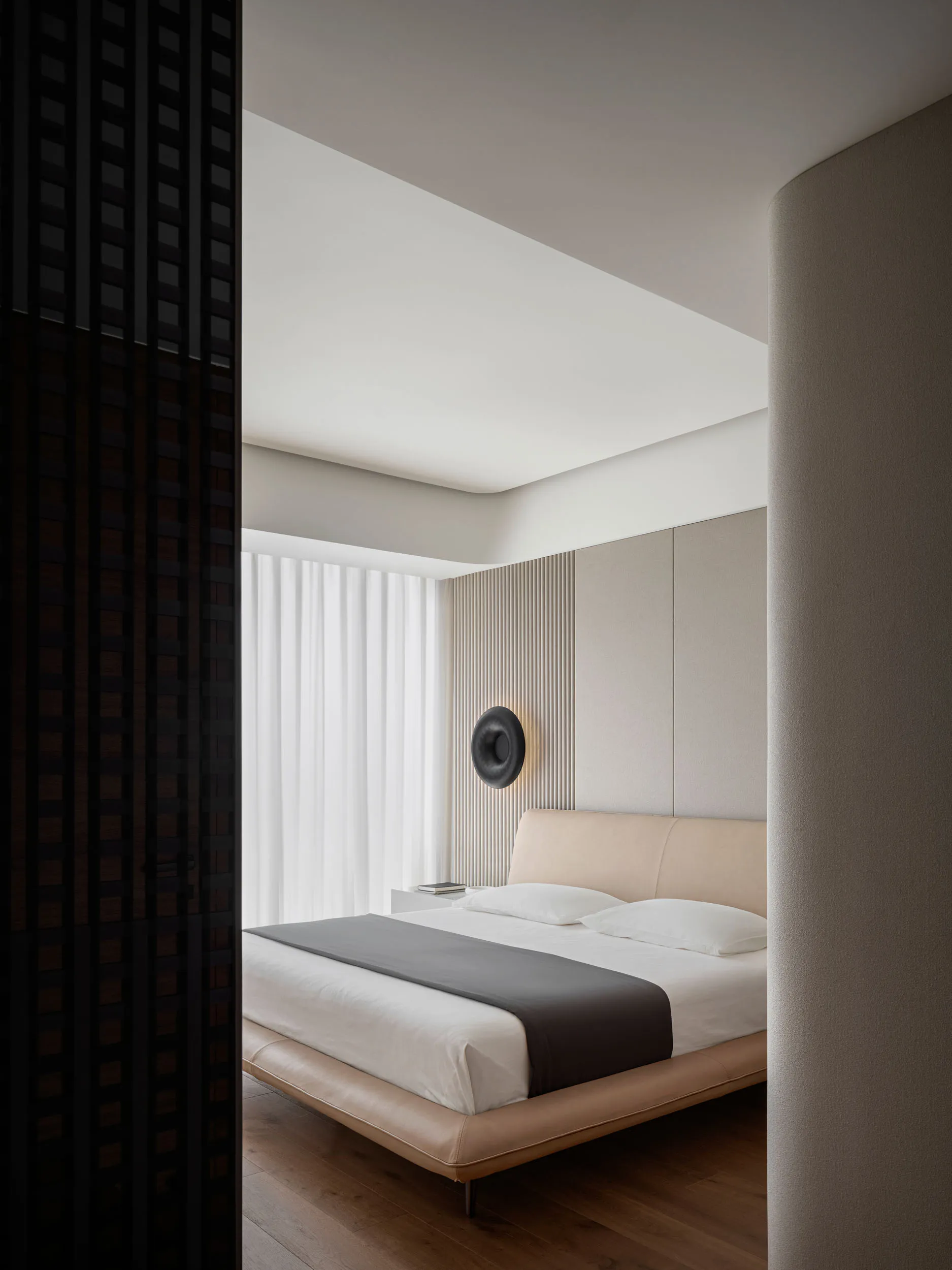 A minimalist bedroom with a beige upholstered bed, wooden flooring, and a round sconce light on the wall.