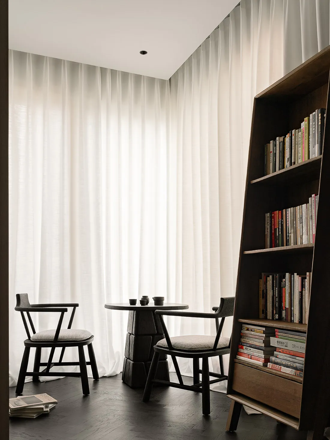 A minimalist residential space featuring wood and glass materials, with a small table and chairs, and a bookshelf.