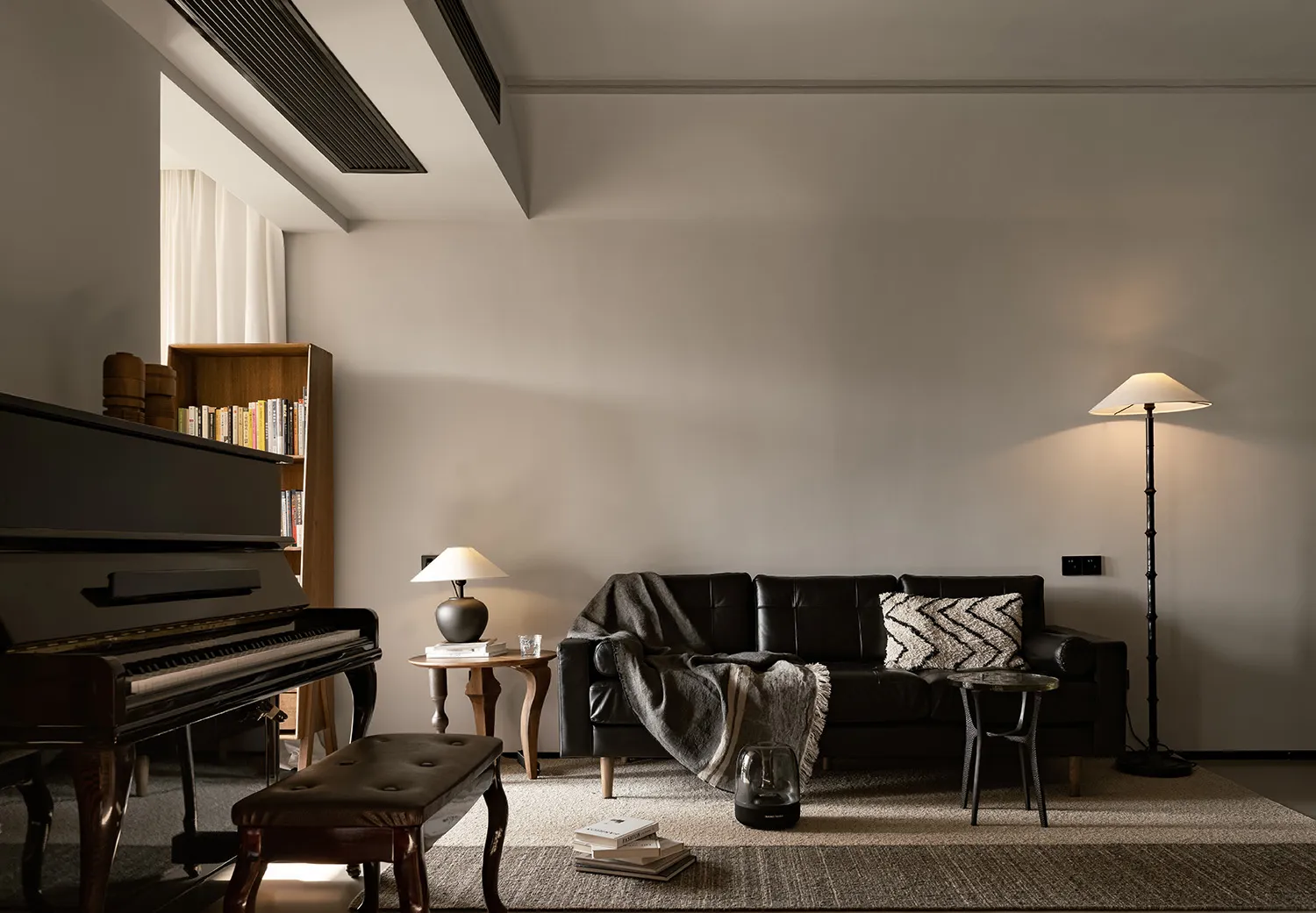 A modern living room featuring a black leather sofa, a floor lamp, and a piano with books and a lamp on a side table.