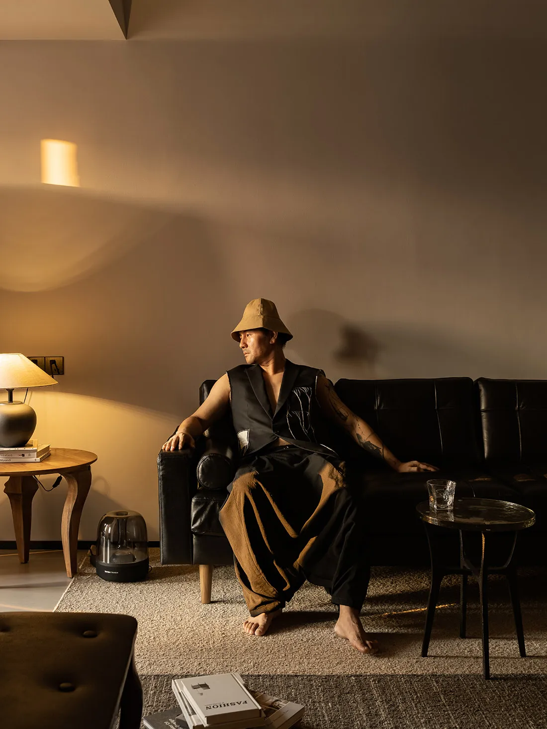 A man wearing a tan bucket hat and a black shirt with a white patch is sitting on a black leather sofa in a living room with