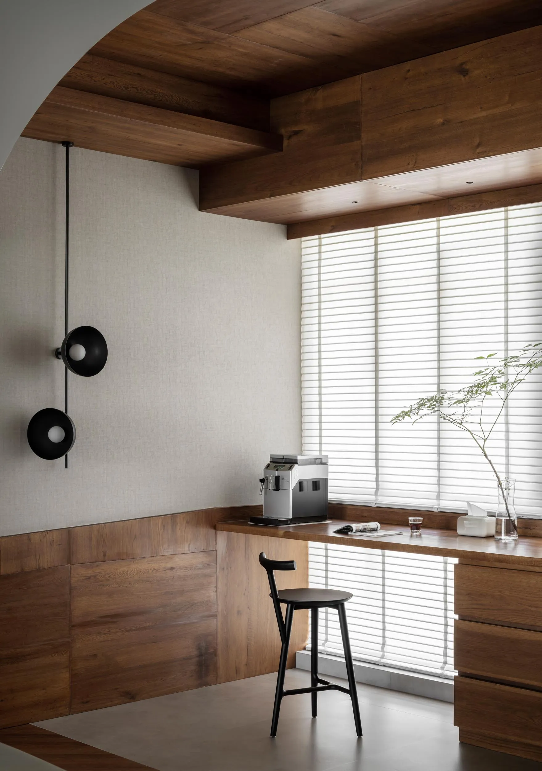 Modern home office with a walnut desk and minimalist design, featuring a black wall lamp and a window with blinds.