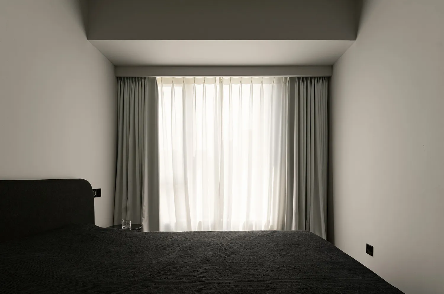 A minimalist bedroom with black furniture and grey walls, featuring white curtains and a window.