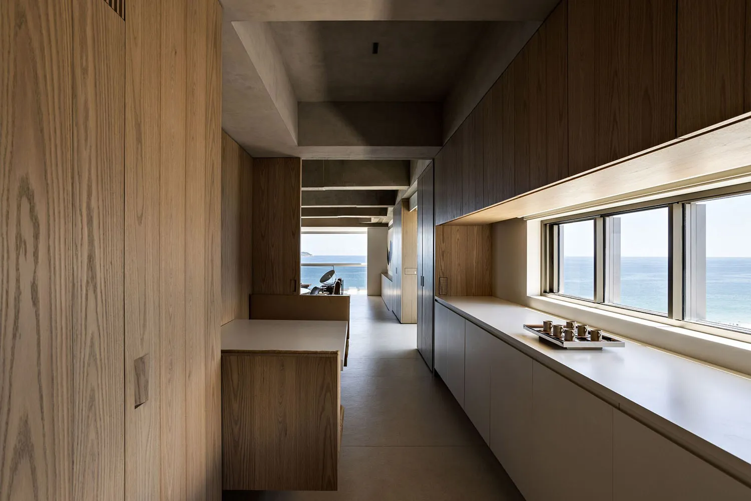 A minimalist modern kitchen with a concrete countertop, wooden cabinets, and a large window overlooking the ocean.