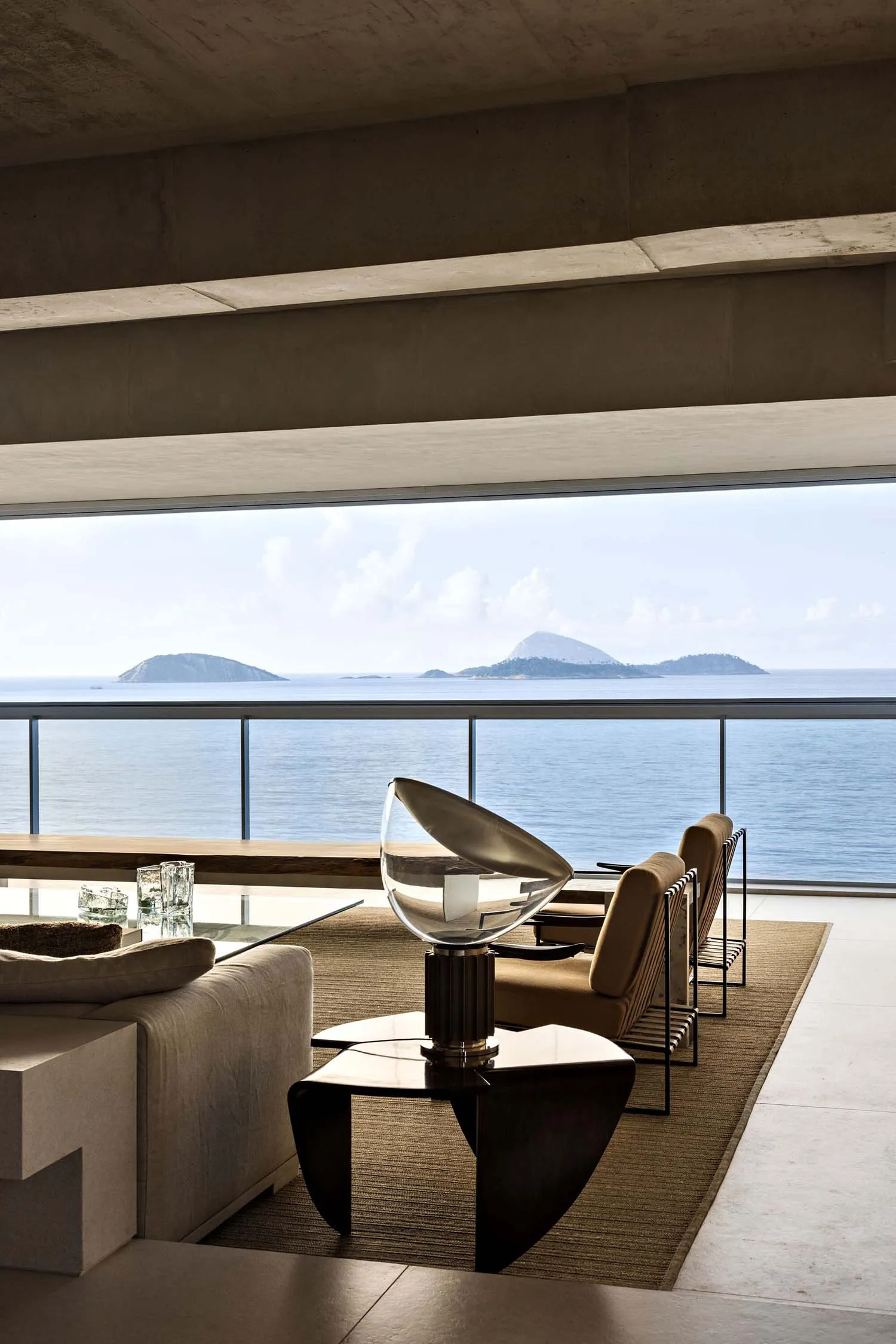 A modern living room with concrete walls, glass furniture, and a view of the ocean.