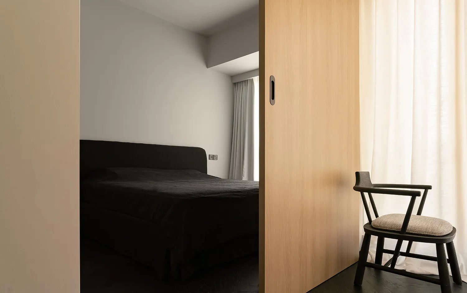 A modern minimalist bedroom with a dark bed, white curtains, and a wooden sliding door.