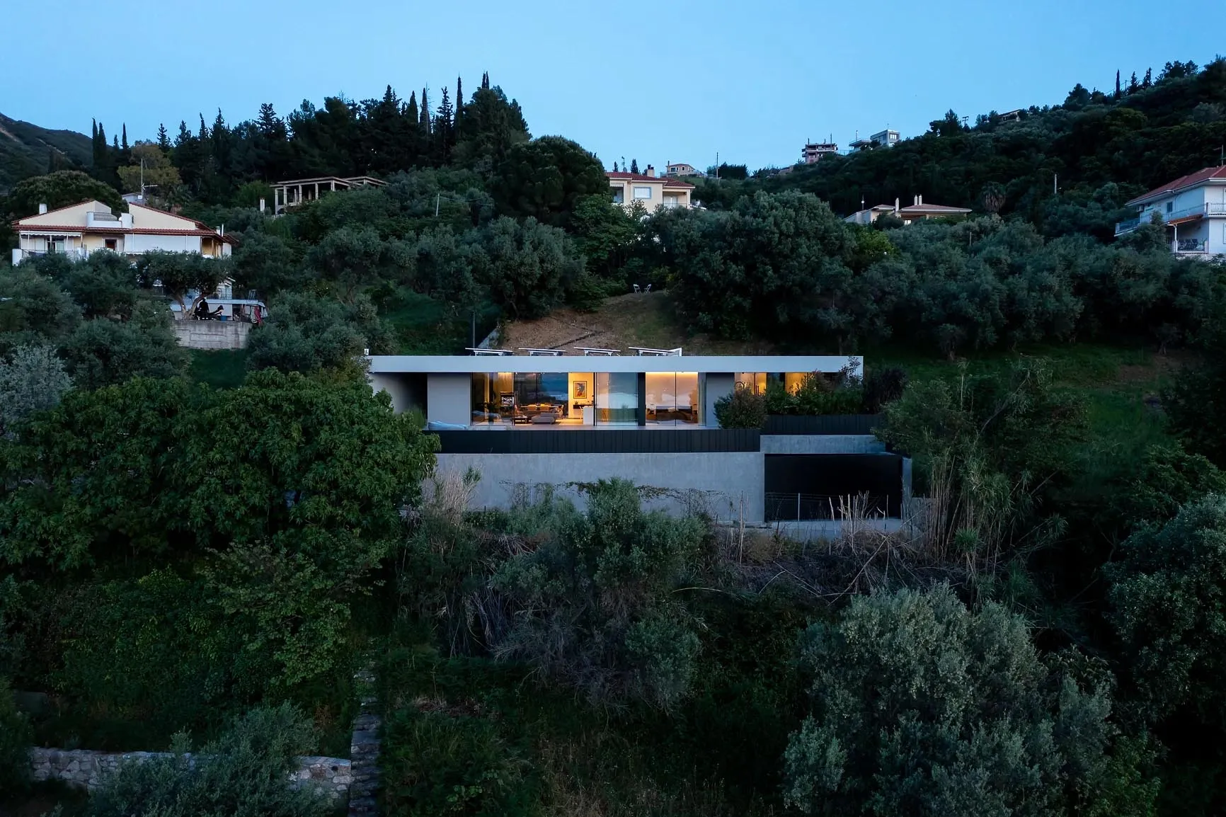 A modern house with concrete walls and large glass windows, surrounded by greenery.