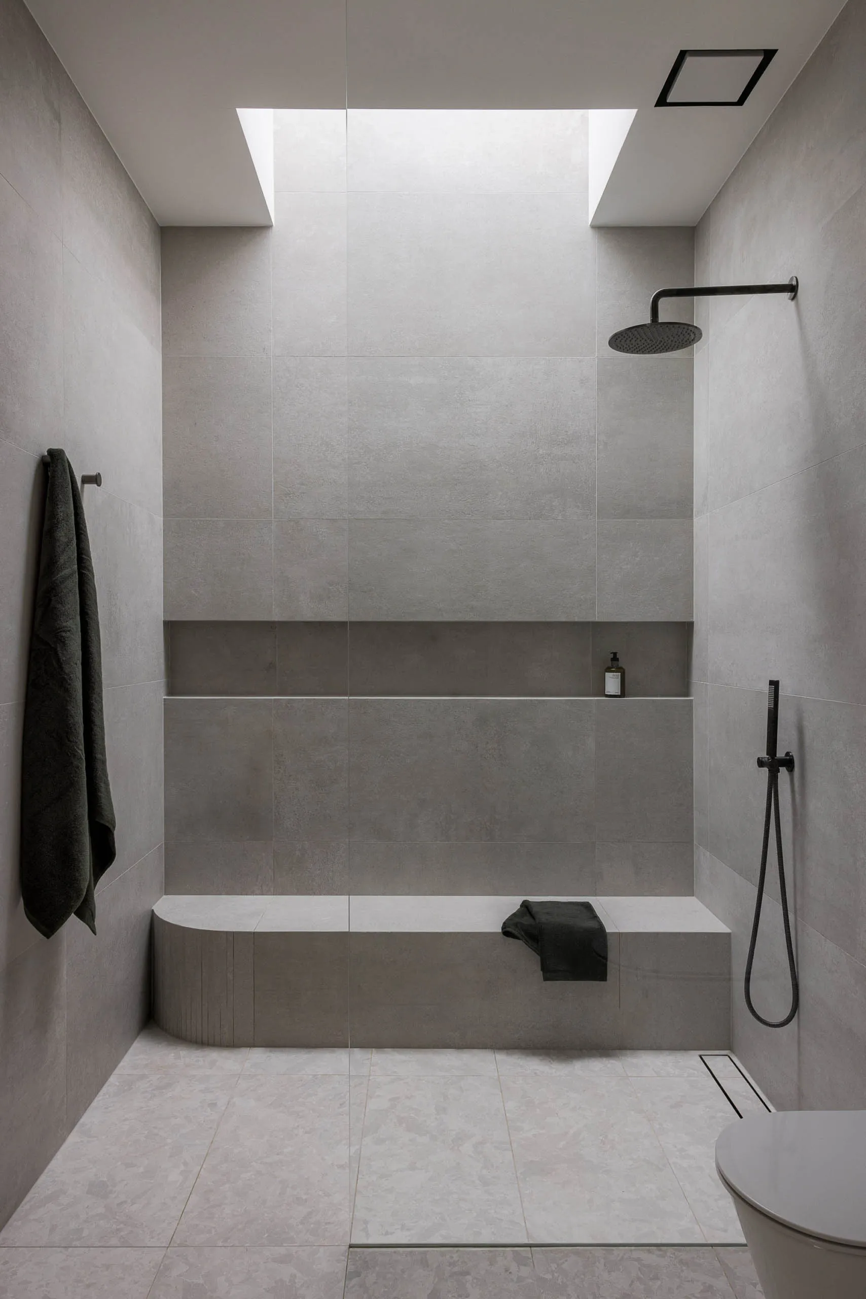Modern minimalist bathroom with concrete walls, large skylight, and built-in bench with a towel hanging nearby.