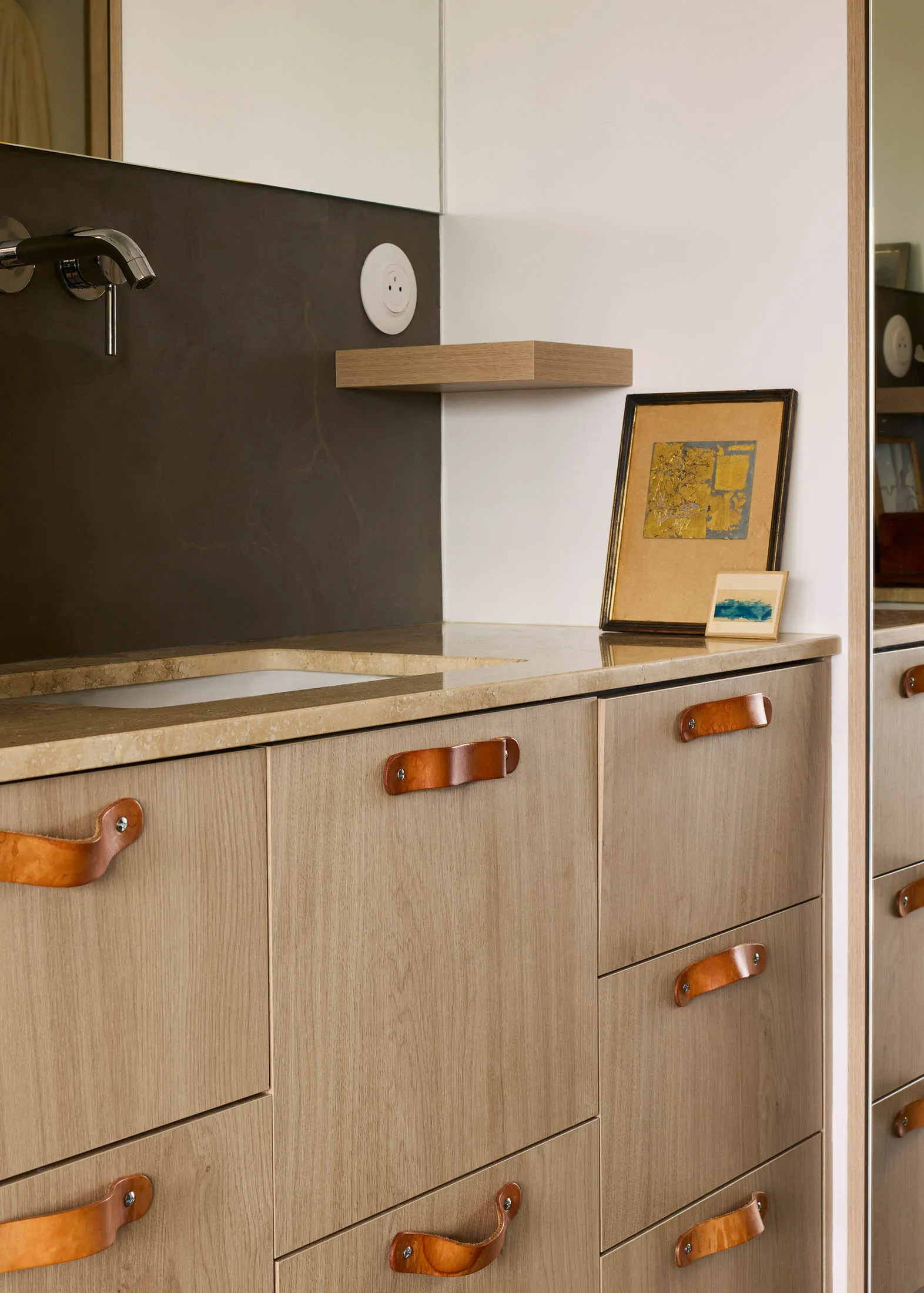 A modern bathroom cabinet featuring a wooden structure with leather handles and a beige marble countertop.