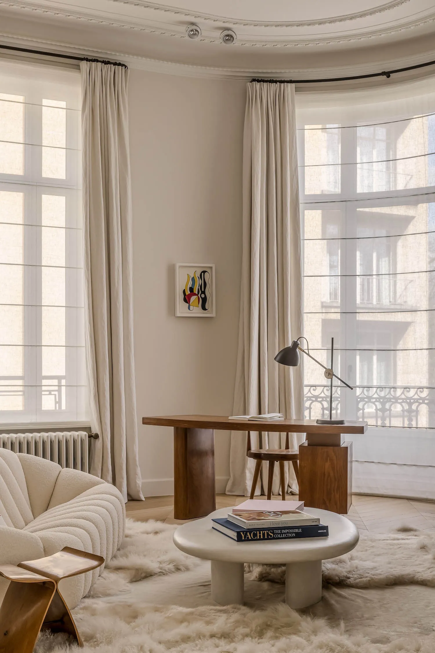A modern living room featuring a white sofa, round table with books, wooden desk with lamp, and large windows with minimalist