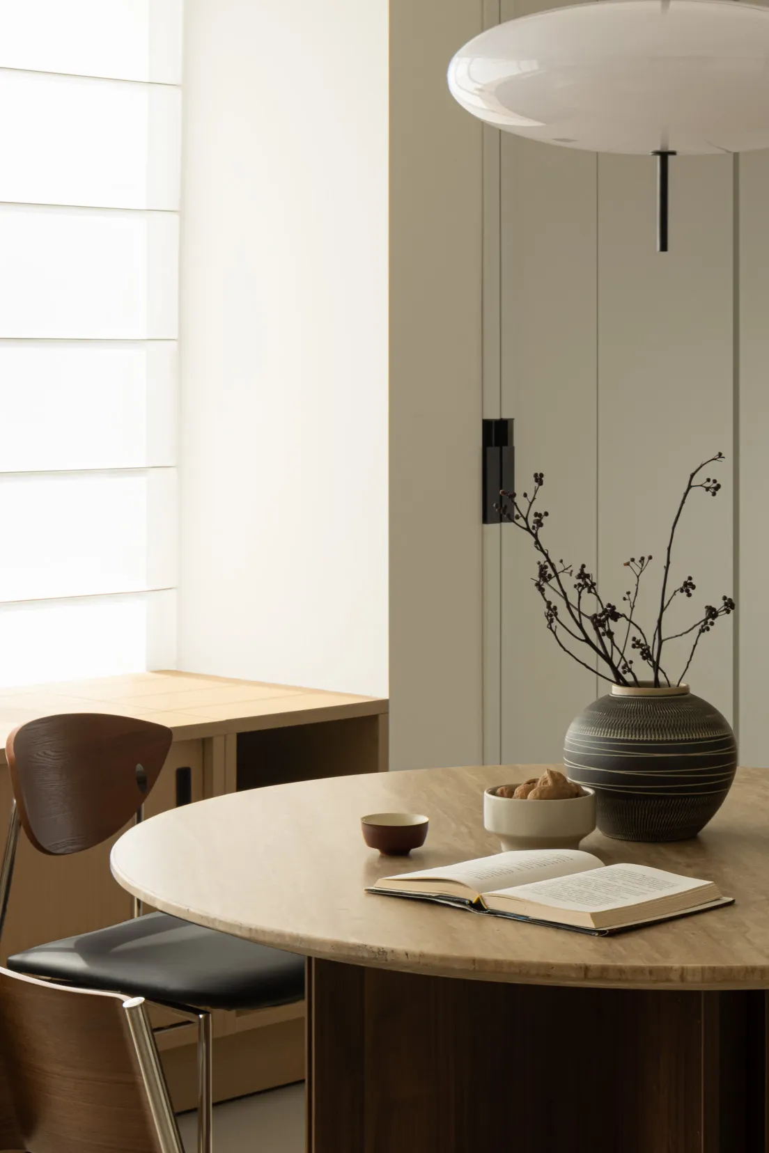 A cozy modern study space featuring wooden furniture, a vase with dried flowers, and an open book on a round wooden table.