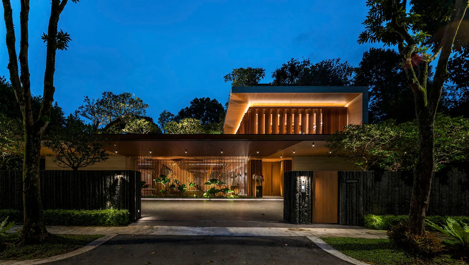Modern entrance with wooden panels of a residential house at dusk, featuring warm lighting and lush greenery.