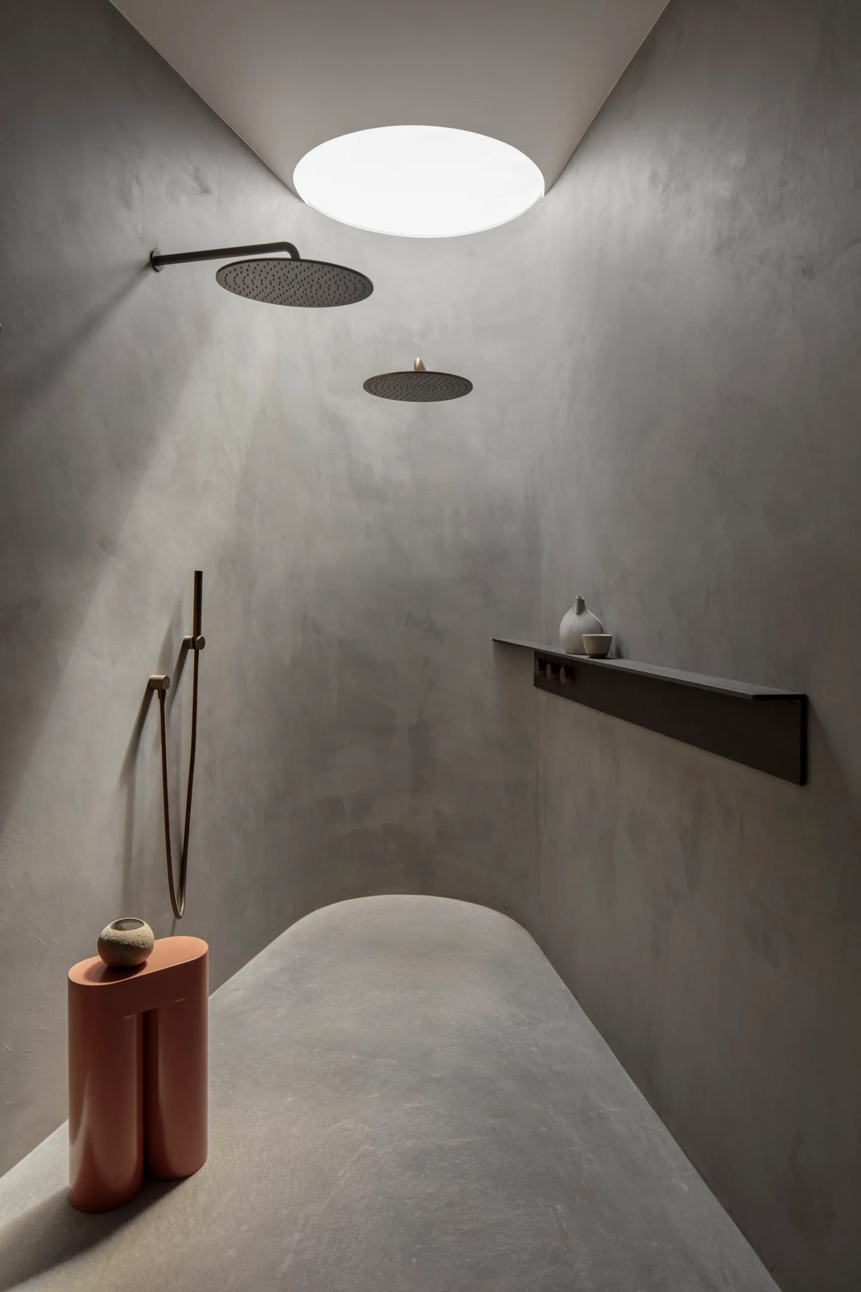 A minimalist bathroom with concrete walls, two overhead showers, and a small shelf with decorative items.