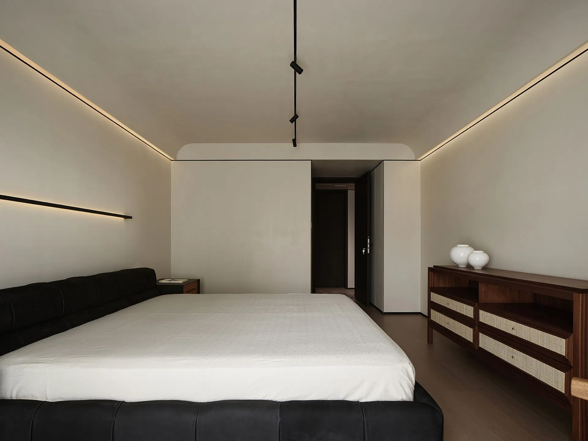 A modern minimalist bedroom features a bed with a dark headboard, white bedding, and neutral-toned walls and flooring.