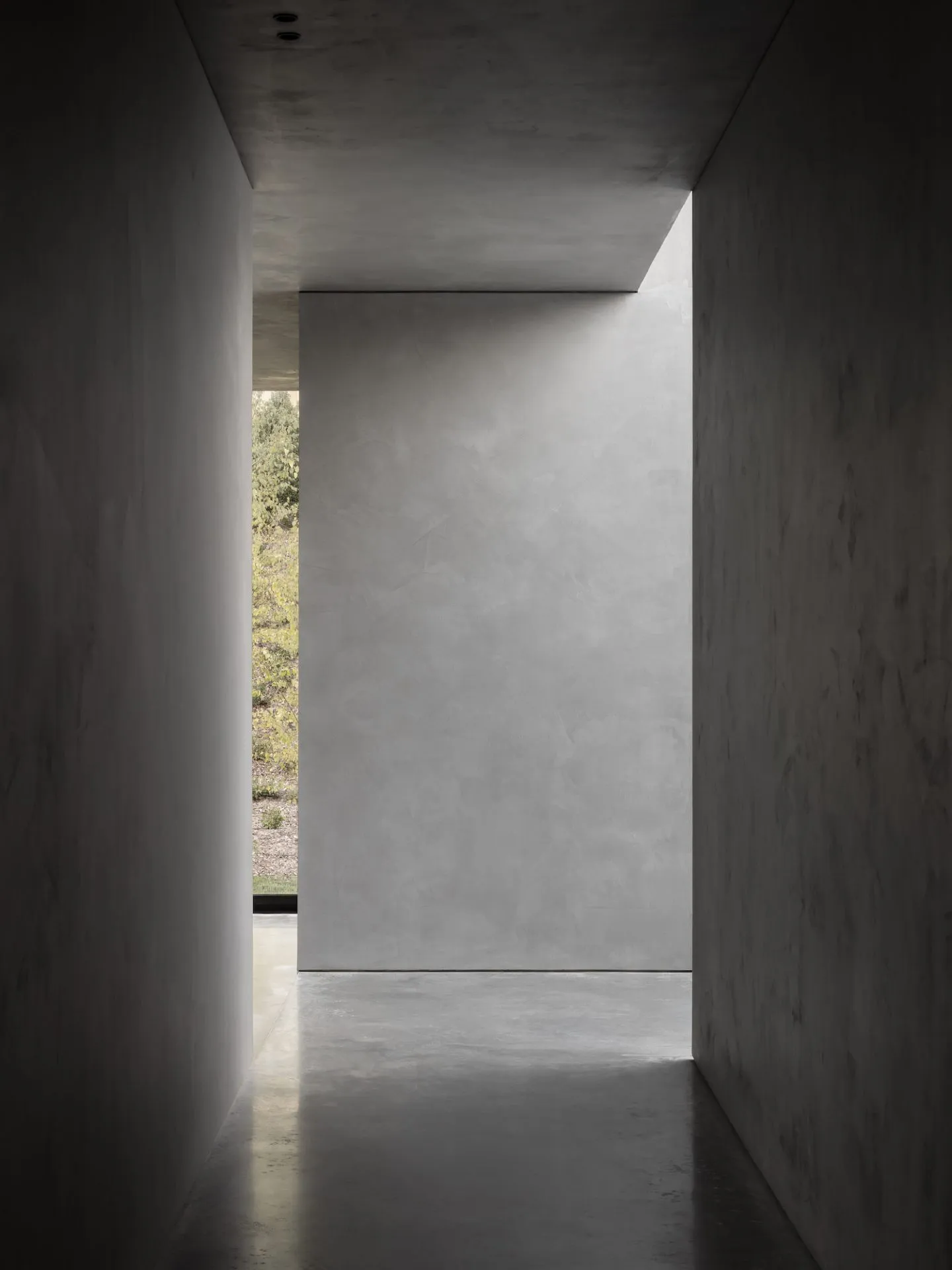 Minimalist interior with concrete walls, floors, and ceilings, leading to a natural light-filled space.