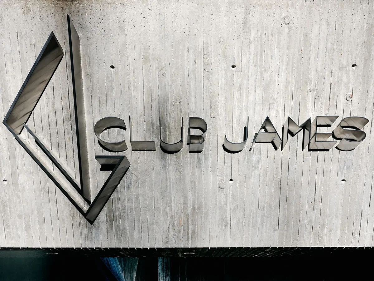 極簡金屬招牌上刻有「CLUB JAMES」字樣,置於混凝土牆背景前。
