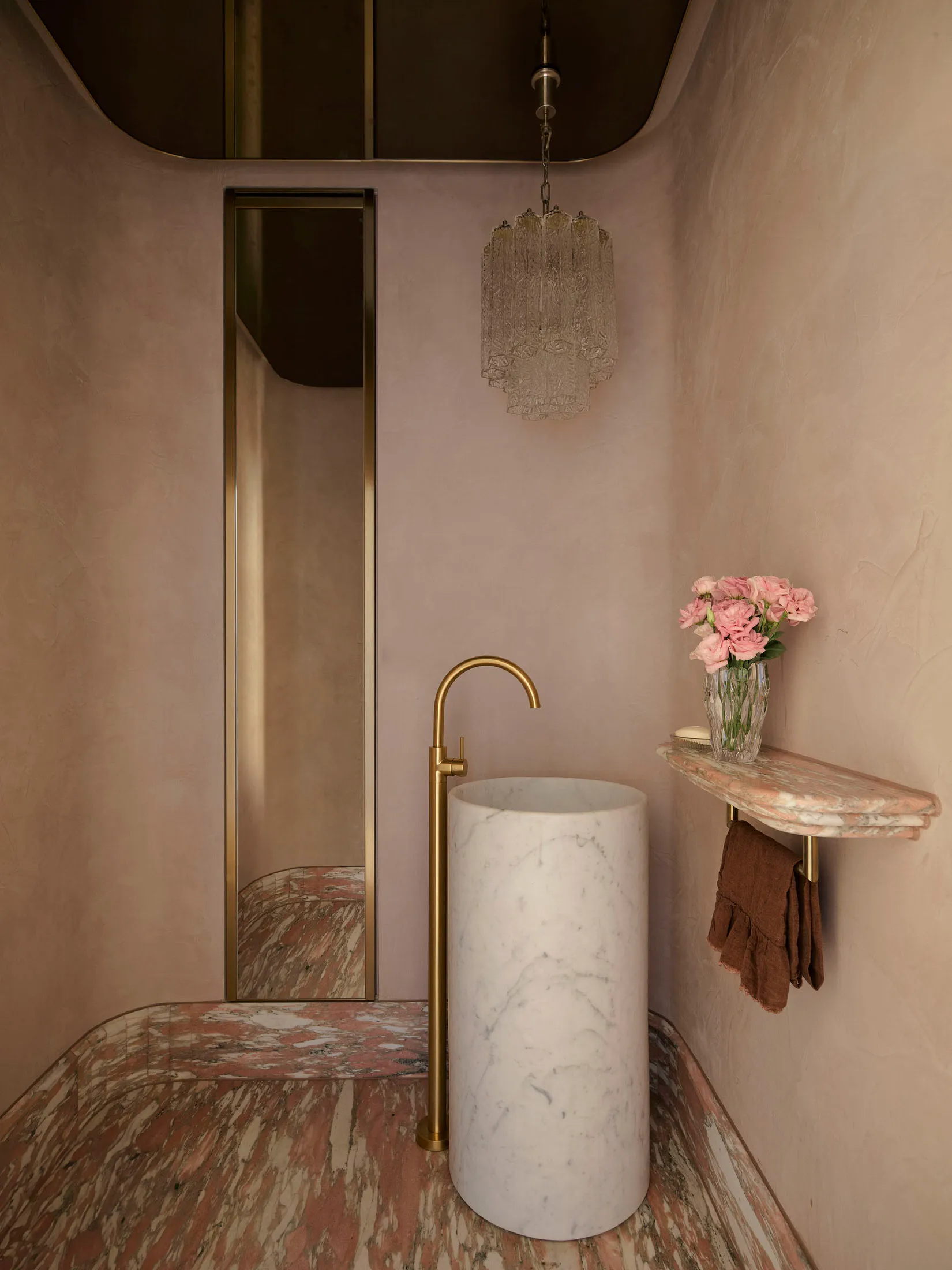 Modern bathroom with marble sink and floor, featuring a brass faucet and a chandelier with pink flowers on the shelf.