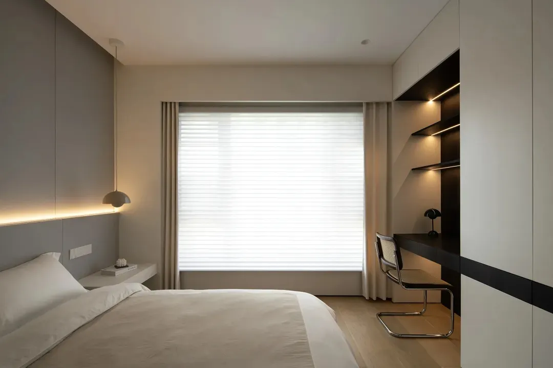 A minimalist bedroom featuring a taupe bed, a built-in desk, and neutral-toned curtains.