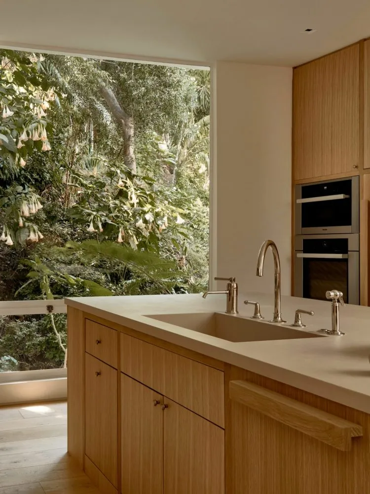 A modern kitchen with wooden cabinets and a large window overlooking a lush green garden.