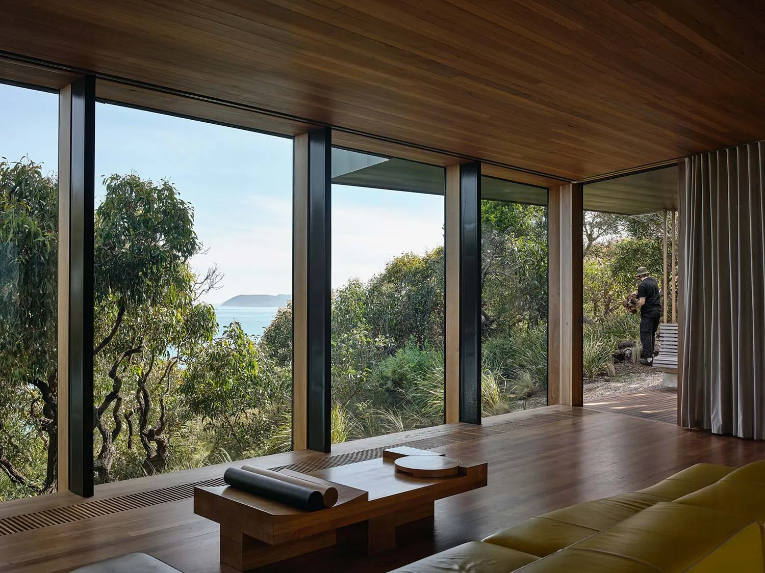 A minimalist living room with large windows overlooking a serene coastal landscape, featuring wooden furniture and a yellow s