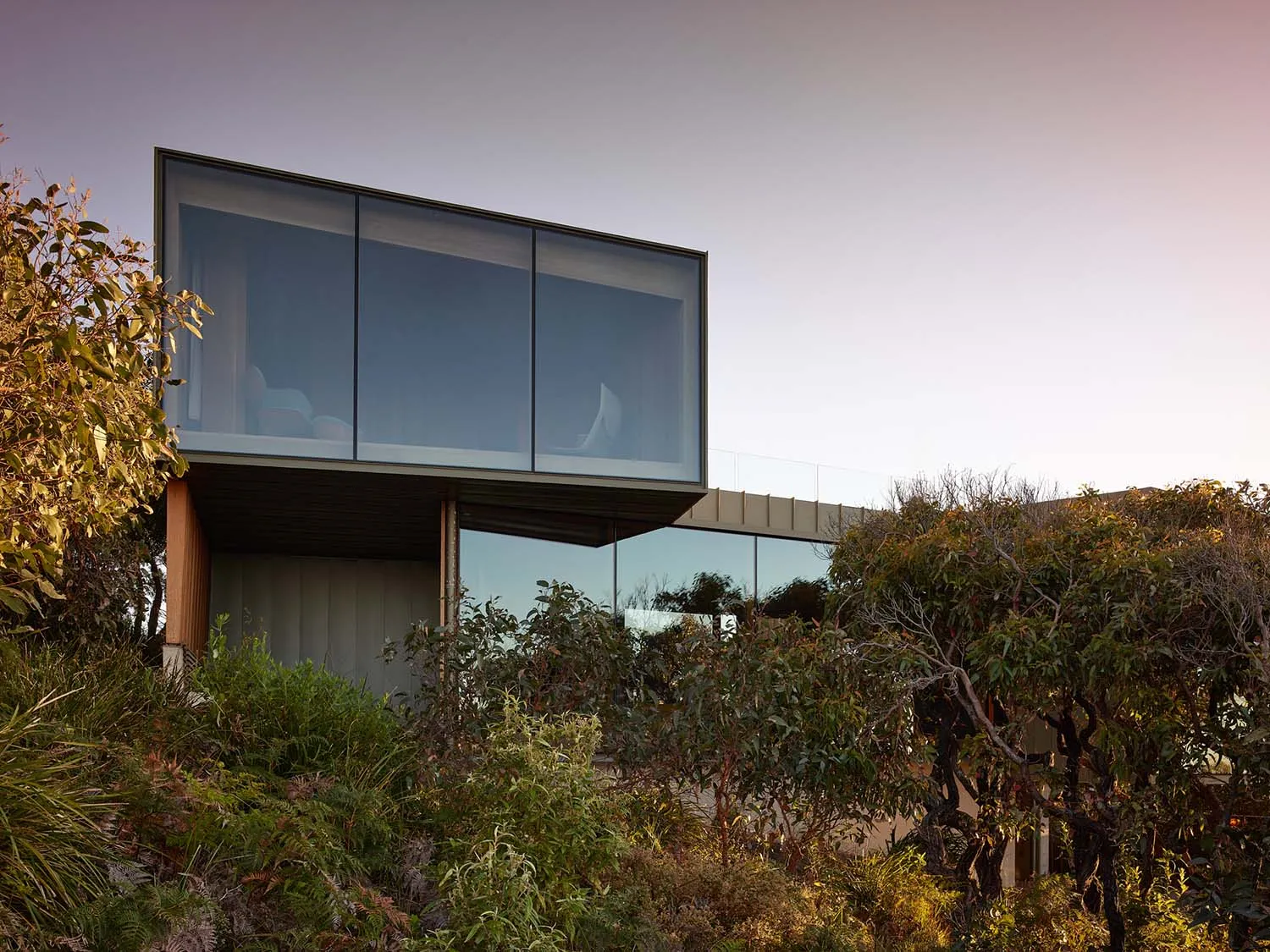 A modern house with transparent glass walls surrounded by greenery.
