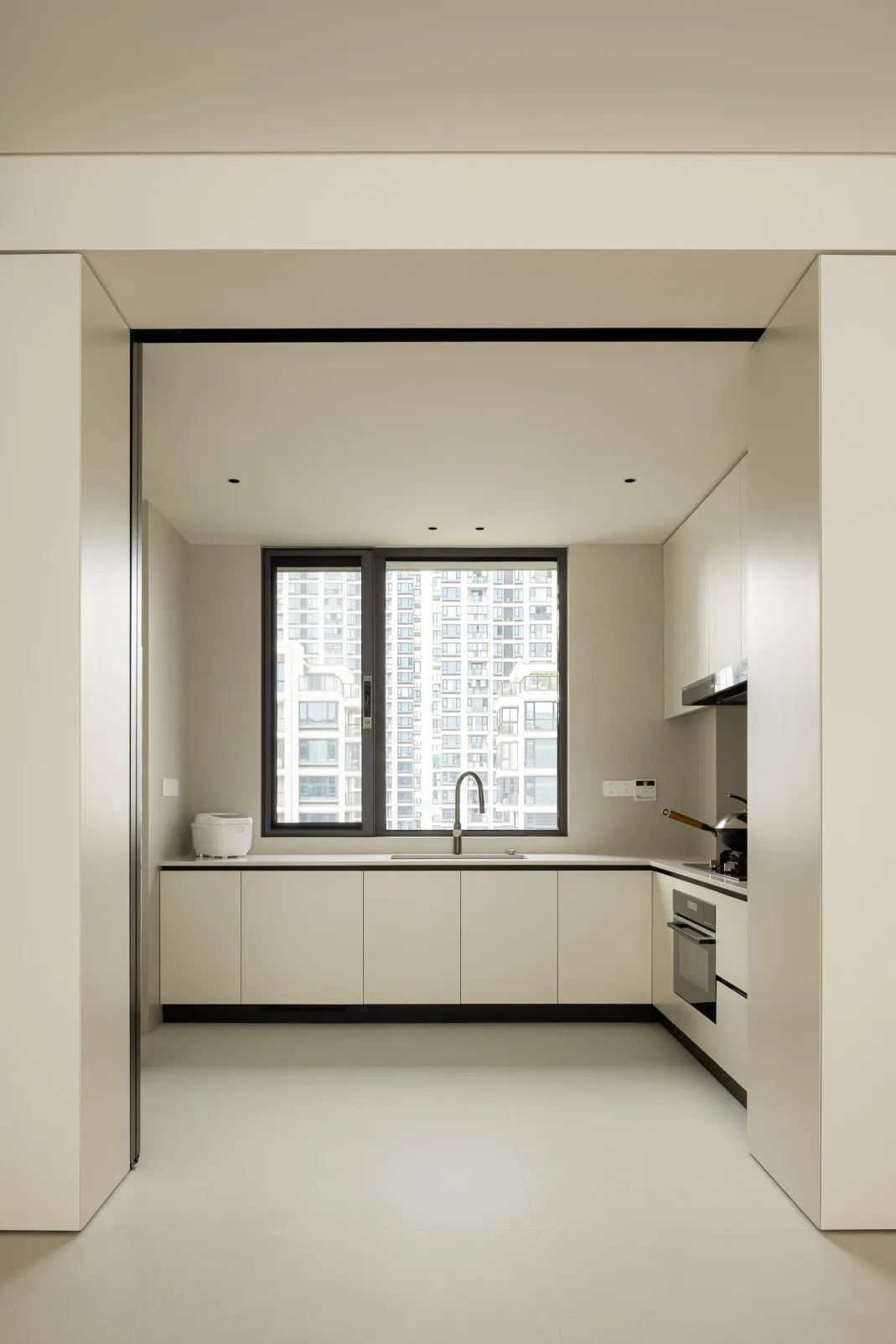 A modern minimalist kitchen with white cabinets and a large window overlooking urban buildings.