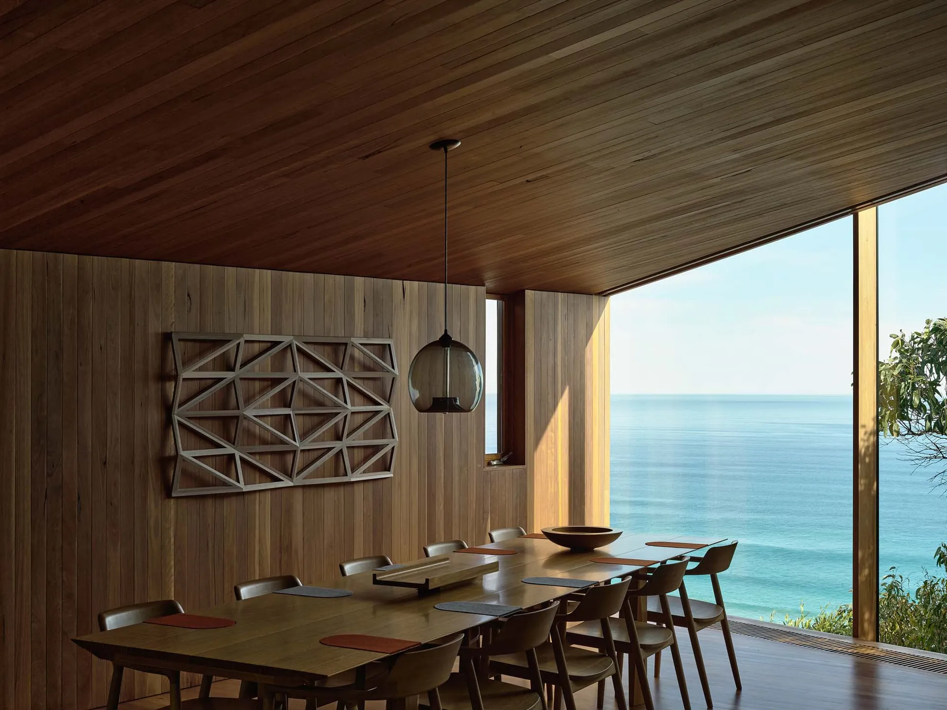 Modern minimalist living room with expansive wooden walls and floor, large windows overlooking the ocean, and a long wooden d