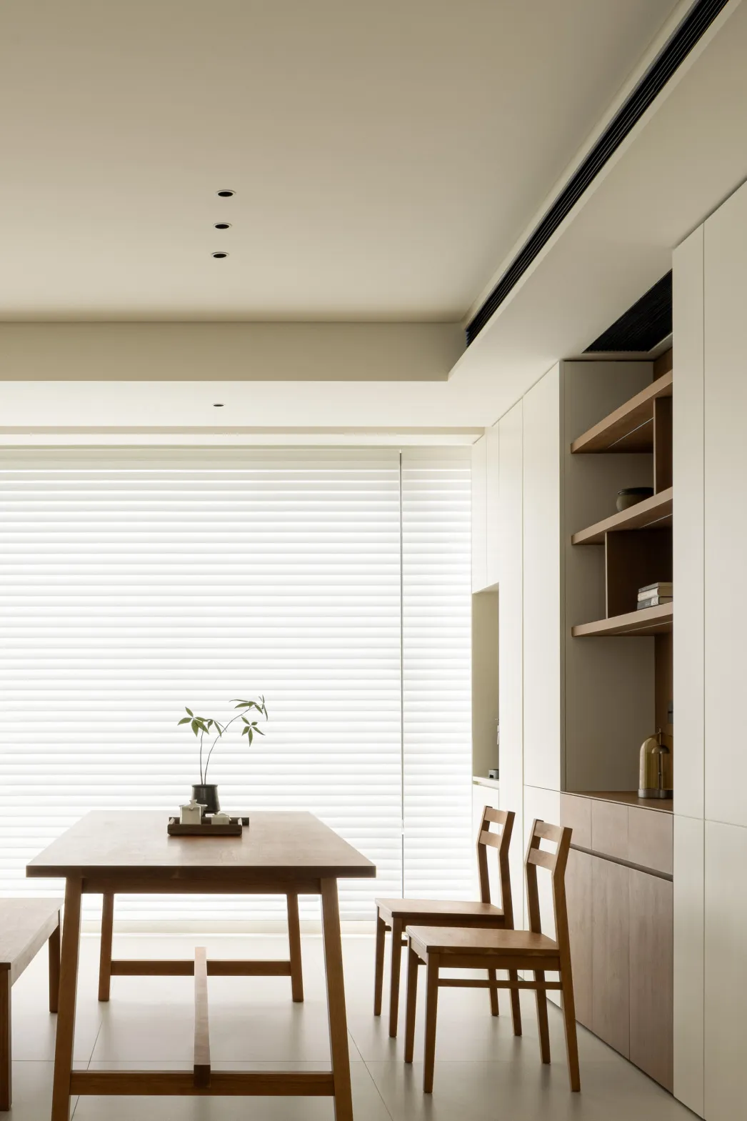 Minimalist dining room featuring a wooden table and chairs with a simple plant centerpiece, against a large window with white