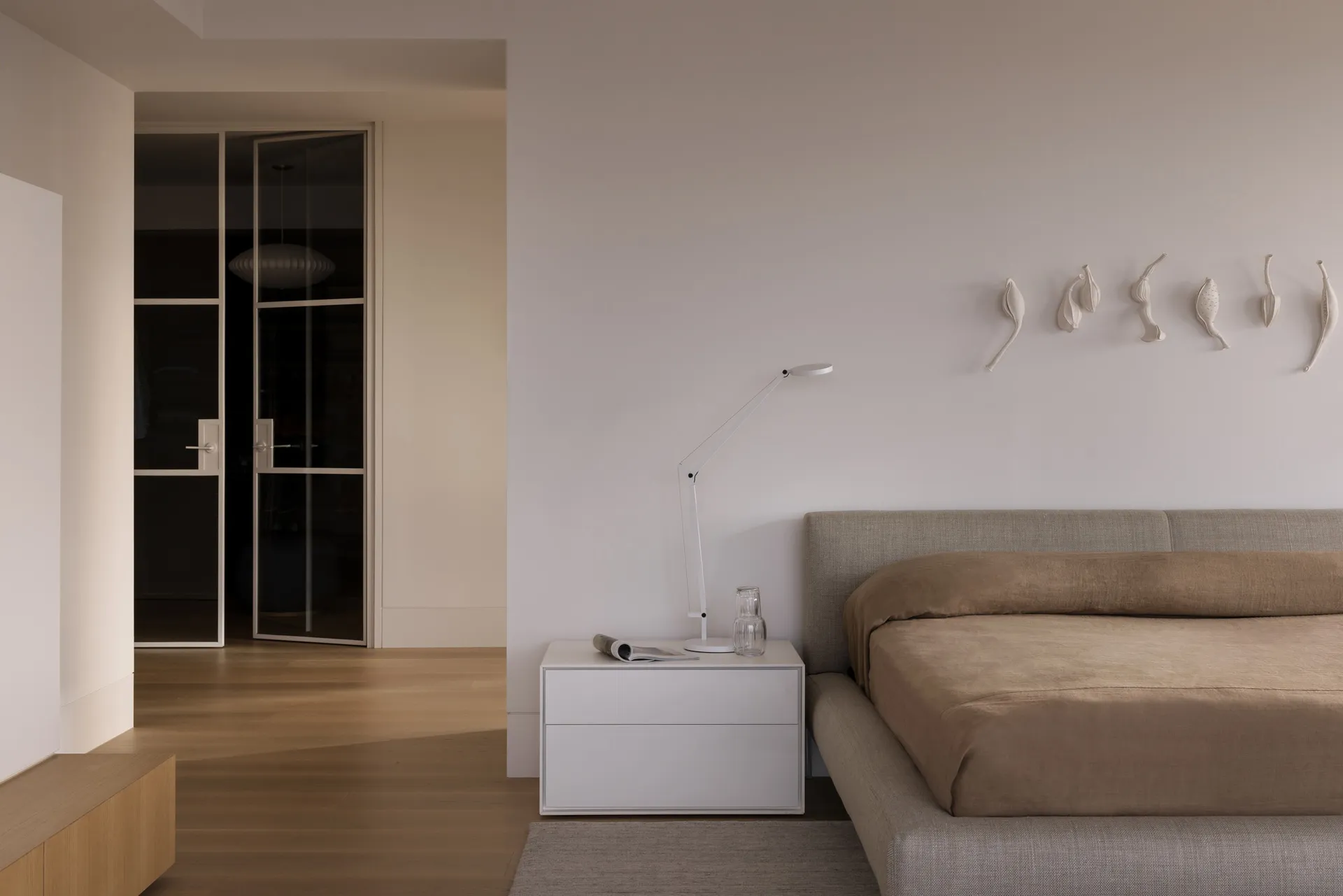 A minimalist bedroom with neutral-toned walls, furniture, and bedding, featuring a simple bed and a white lamp on a white sid