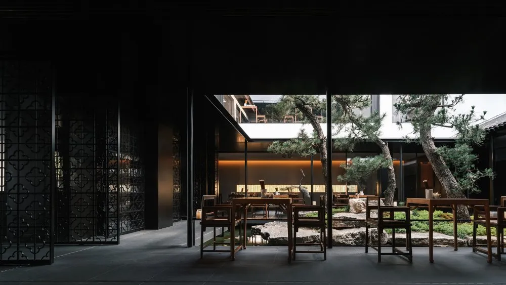 Modern restaurant courtyard with wooden tables, glass walls, and stone elements.