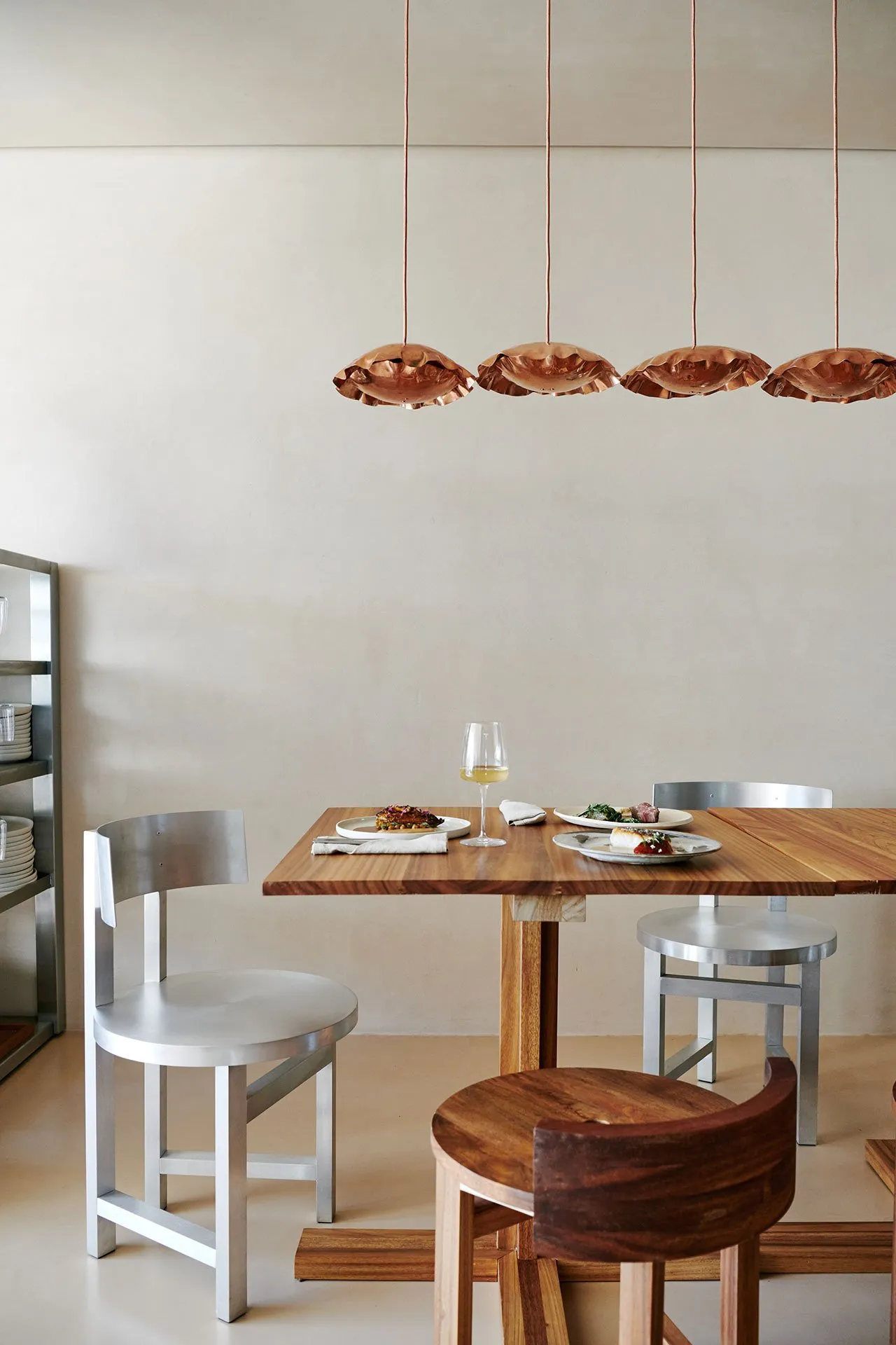 A minimalist dining room featuring a wooden table and metal chairs under a copper pendant light fixture.