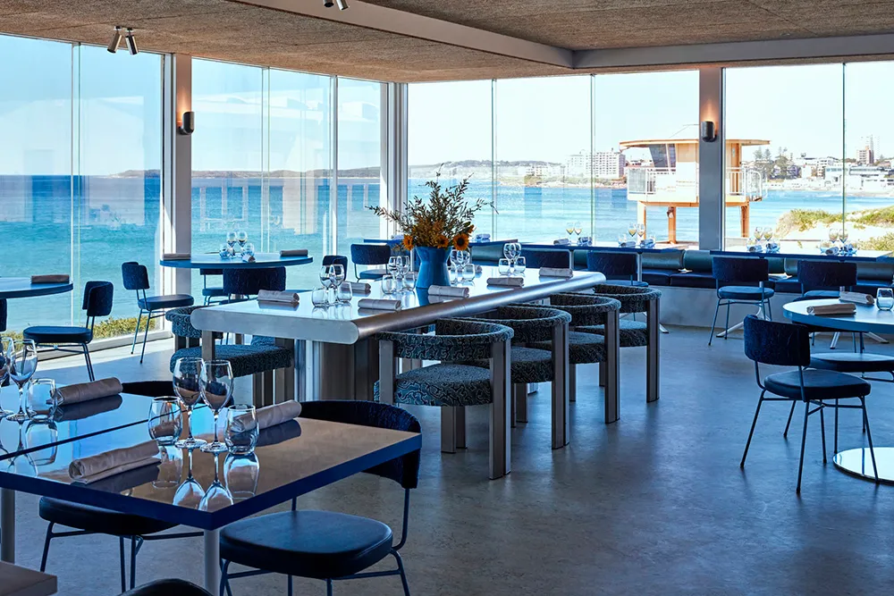 Modern restaurant interior with glass walls and tables overlooking the ocean.