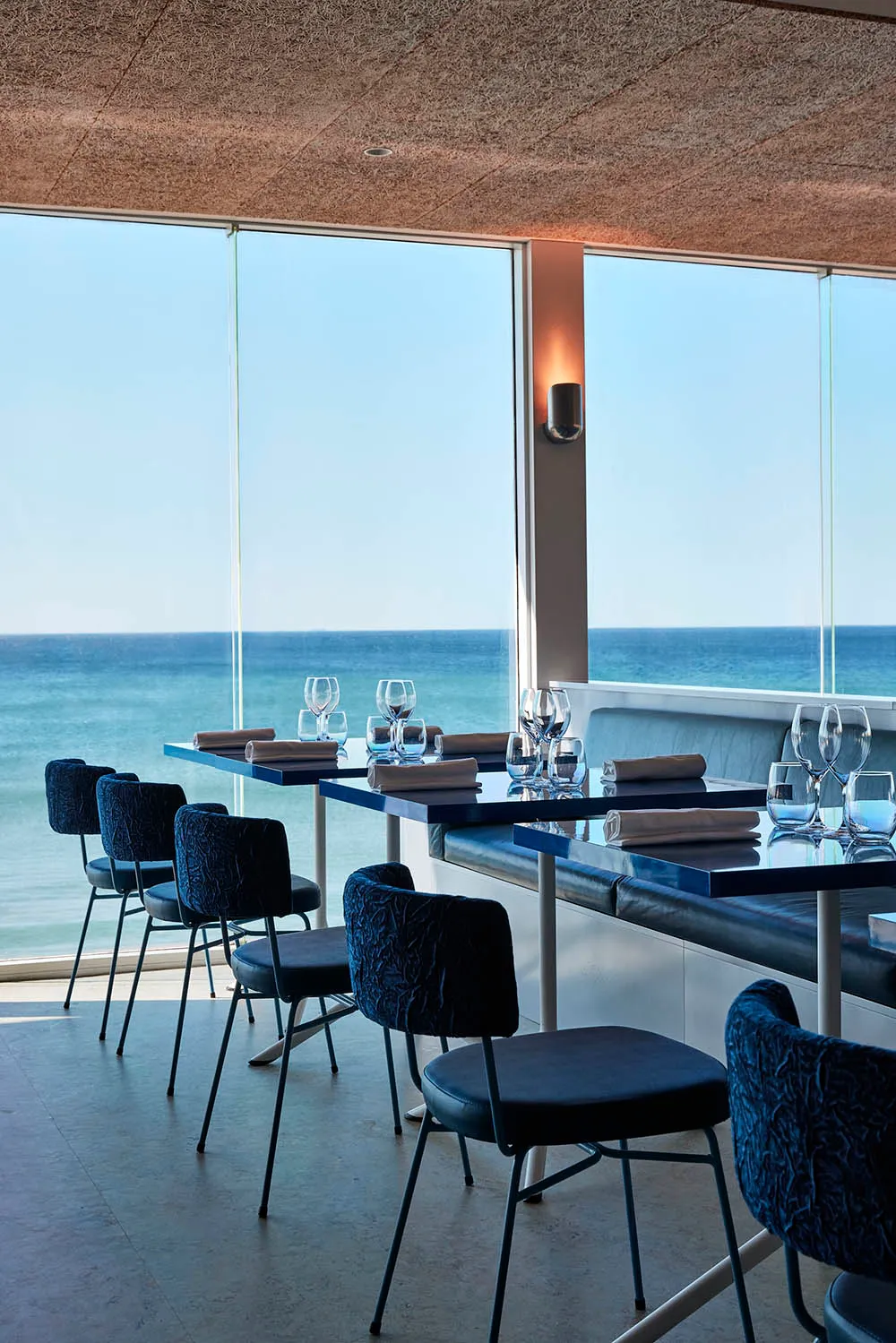 A modern restaurant interior featuring tables, chairs, and wine glasses with a glass wall overlooking the ocean.