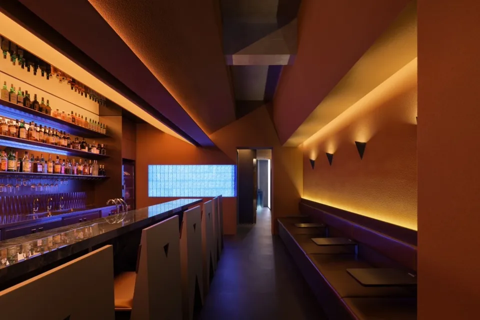 A modern industrial-style bar interior with concrete walls, shelves of bottles, and seating.