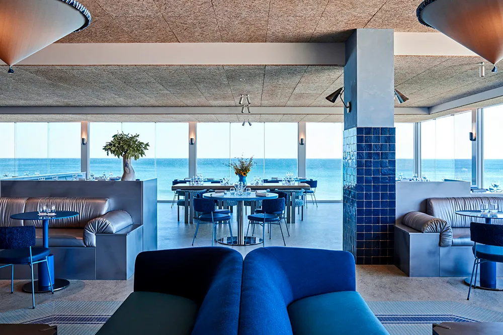 A modern restaurant interior with sea views, featuring blue sofas, tables, chairs, and decorative vases.
