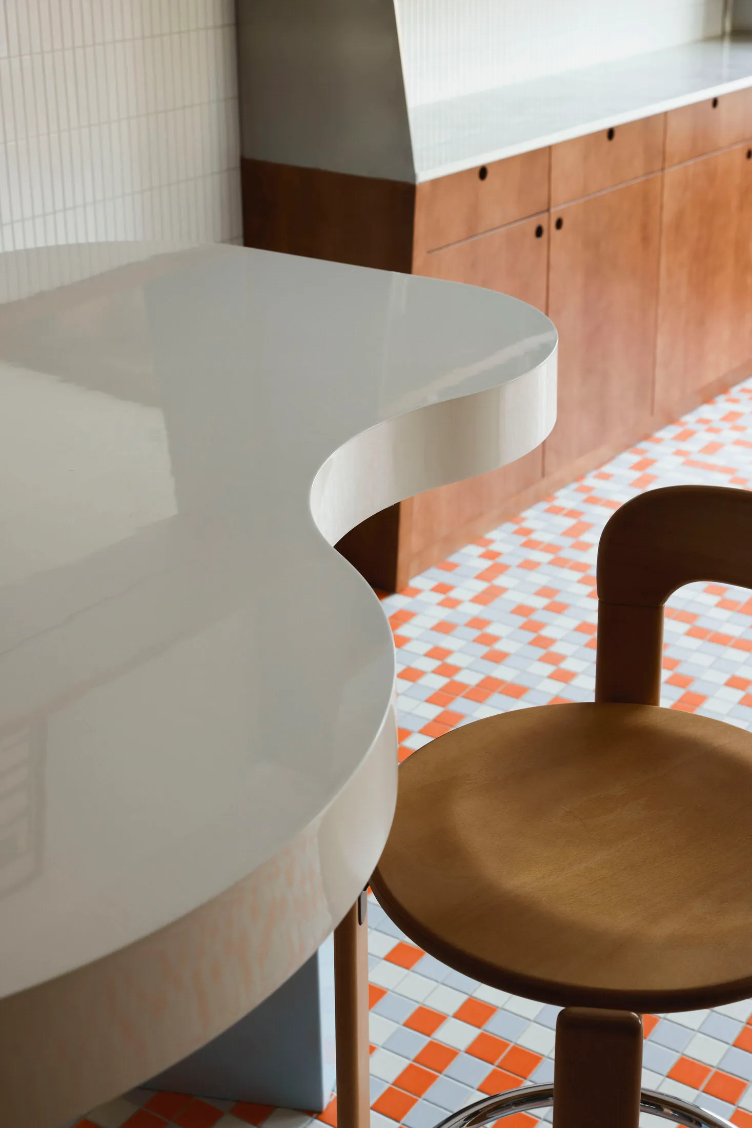 A modern commercial kitchen with curved white countertops, wooden stools, and a mix of white and orange checkered tiles on th