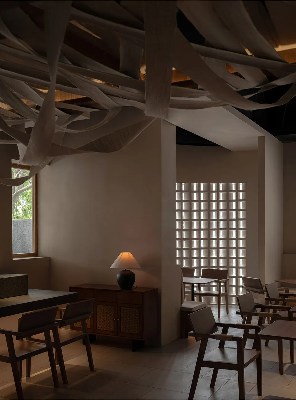 A minimalist restaurant interior featuring wooden tables, chairs, and a wooden cabinet with a lamp, with fabric-covered beams