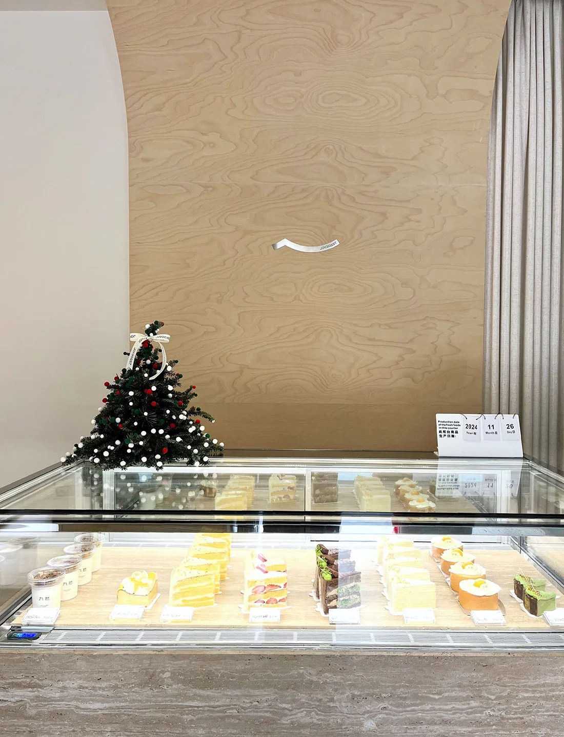 A glass bakery display case featuring various slices of cakes and desserts, adorned with a small, decorated Christmas tree.