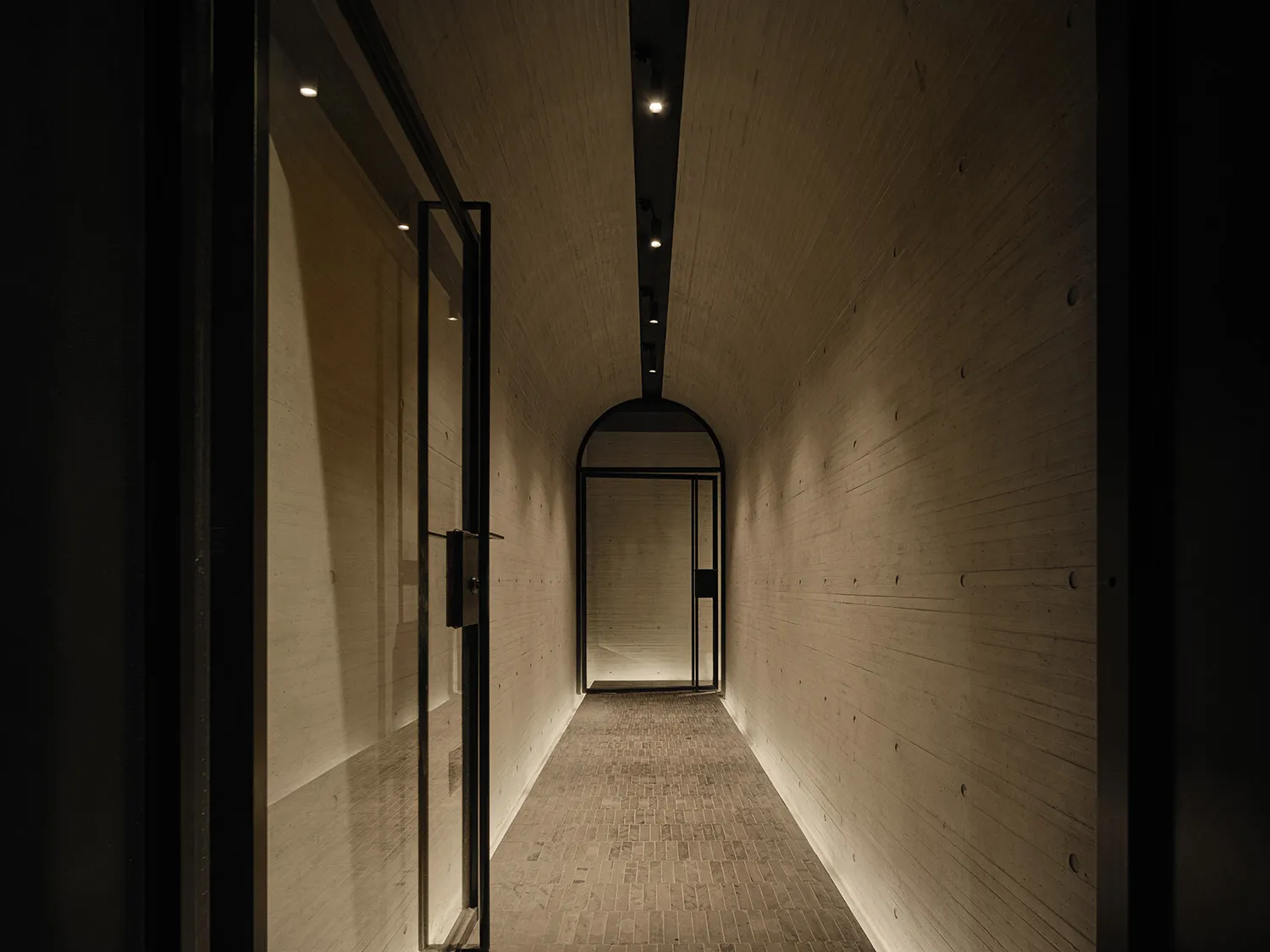 A minimalist corridor with concrete walls and a tiled floor, featuring a single door at the end.