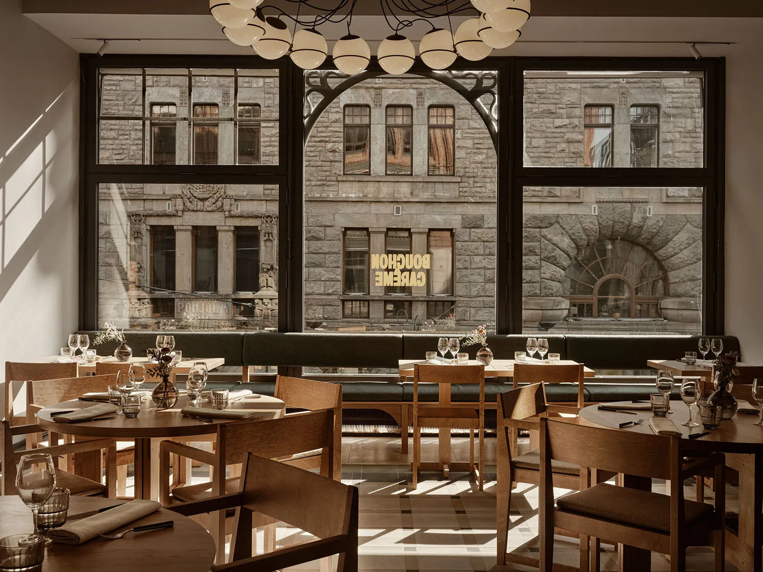 Interior view of a modern cafe featuring wooden seating and large windows overlooking a stone building.