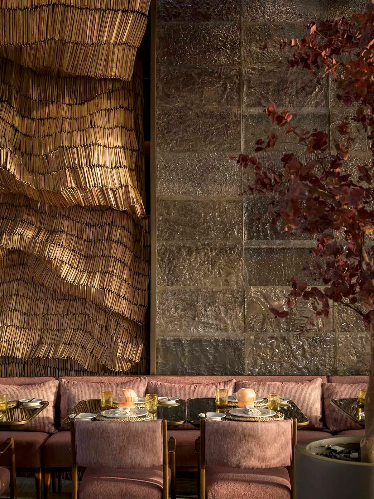 A modern restaurant interior with a woven textile wall and a textured concrete backdrop, featuring brass chairs and tables wi