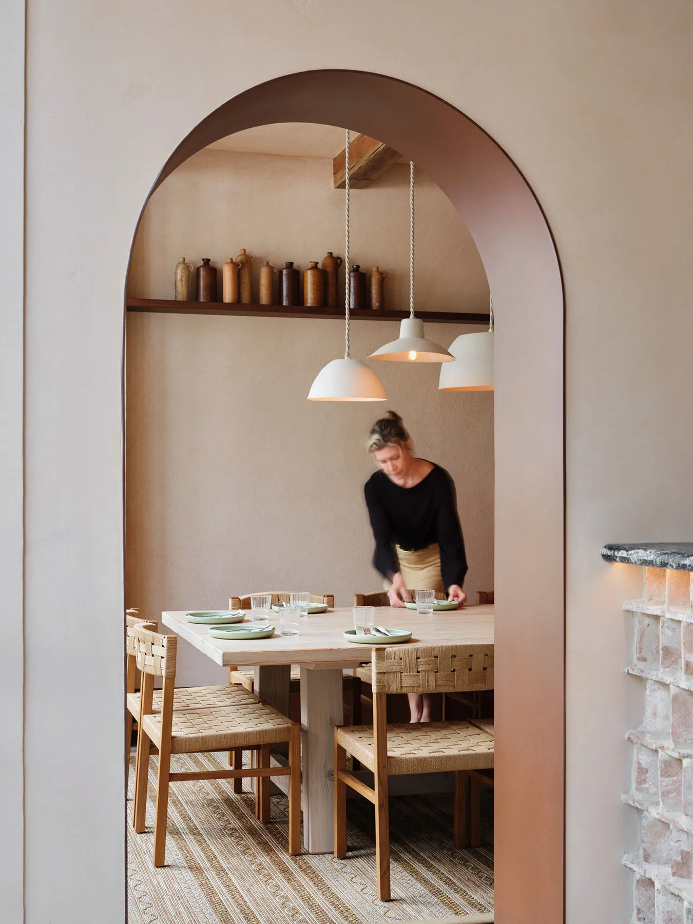 A warm-toned restaurant interior with a wooden dining table, woven chairs, and pendant lights hanging above.
