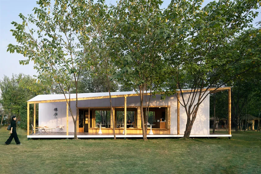A modern residential home featuring glass windows and a wooden frame, surrounded by greenery and a clear sky.