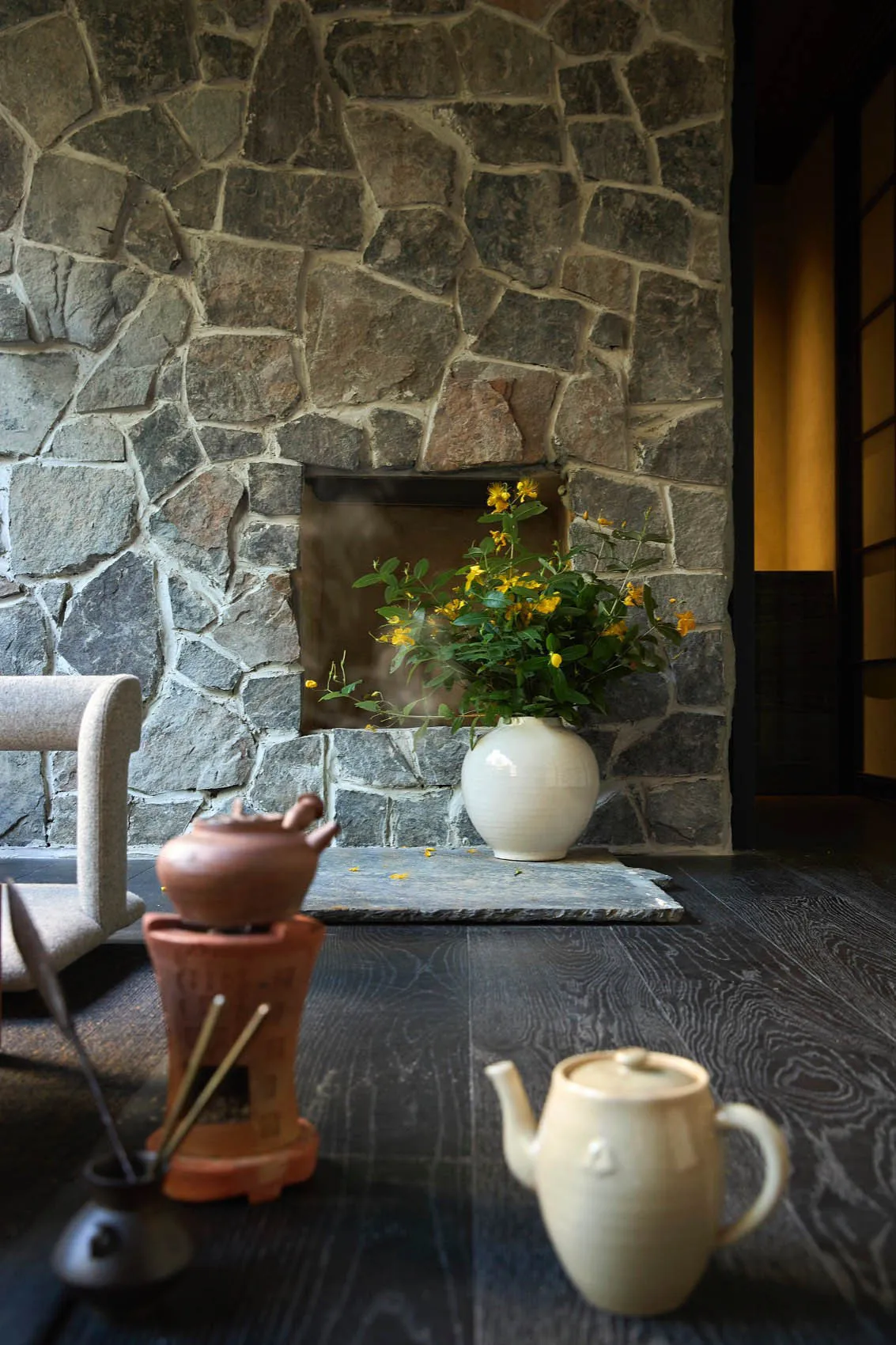 A cozy living room with a stone fireplace, wooden floor, and a white vase with yellow flowers.