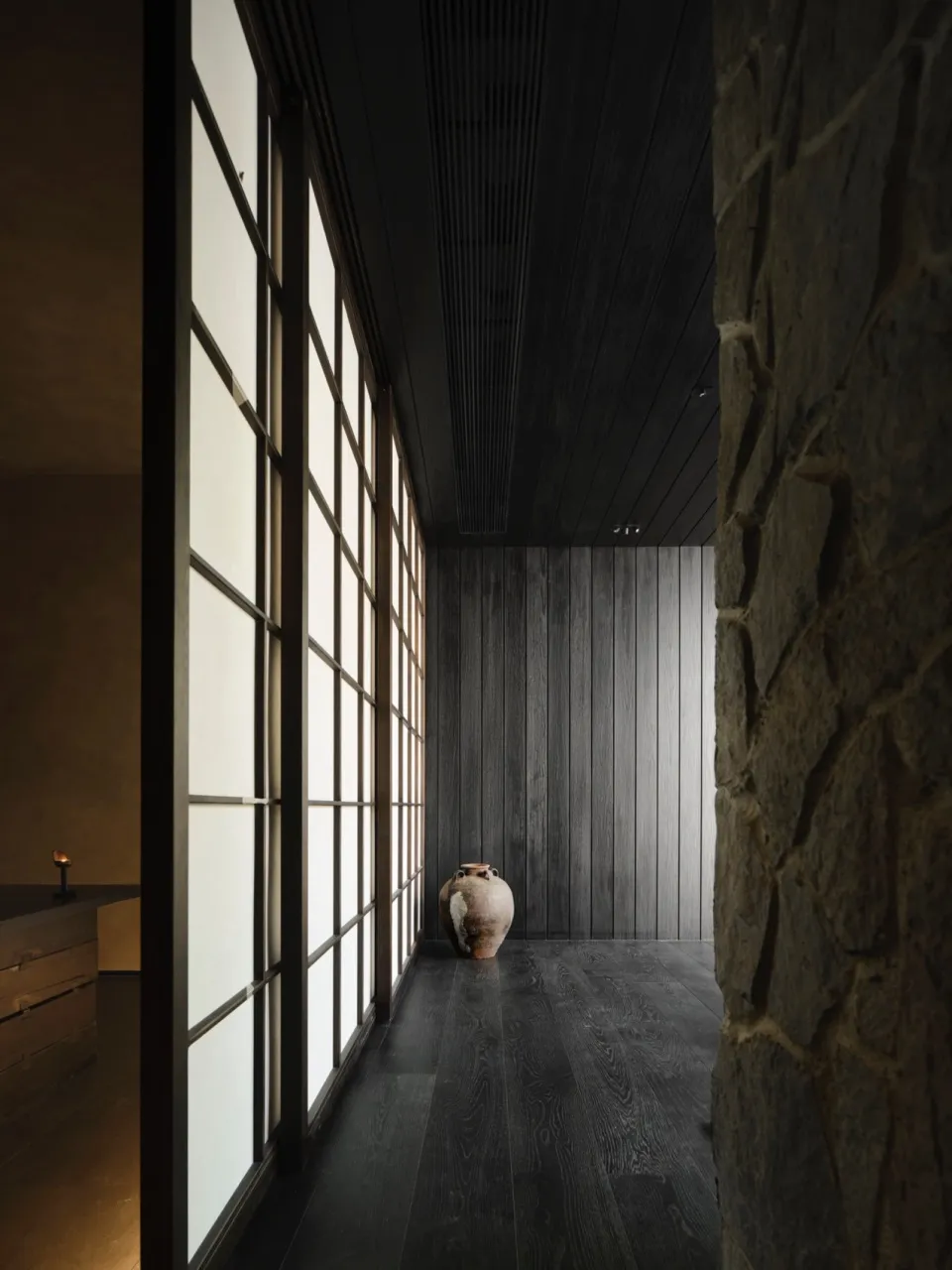 A minimalist modern interior space featuring wood paneling and traditional Japanese shoji screens, with a vase on the floor.