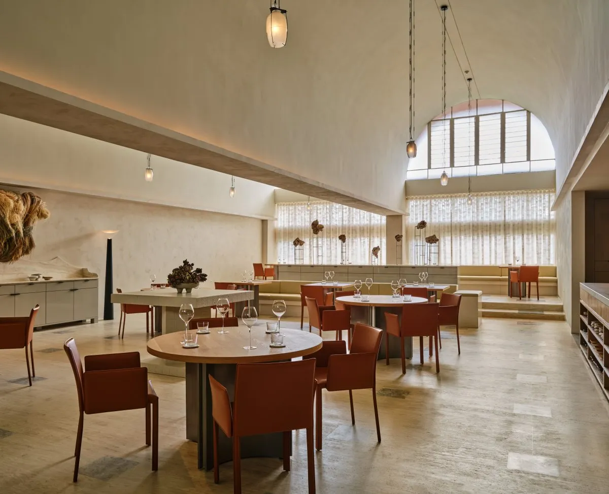 A spacious, modern restaurant interior featuring concrete tables and chairs with high ceilings, pendant lights, and arches.