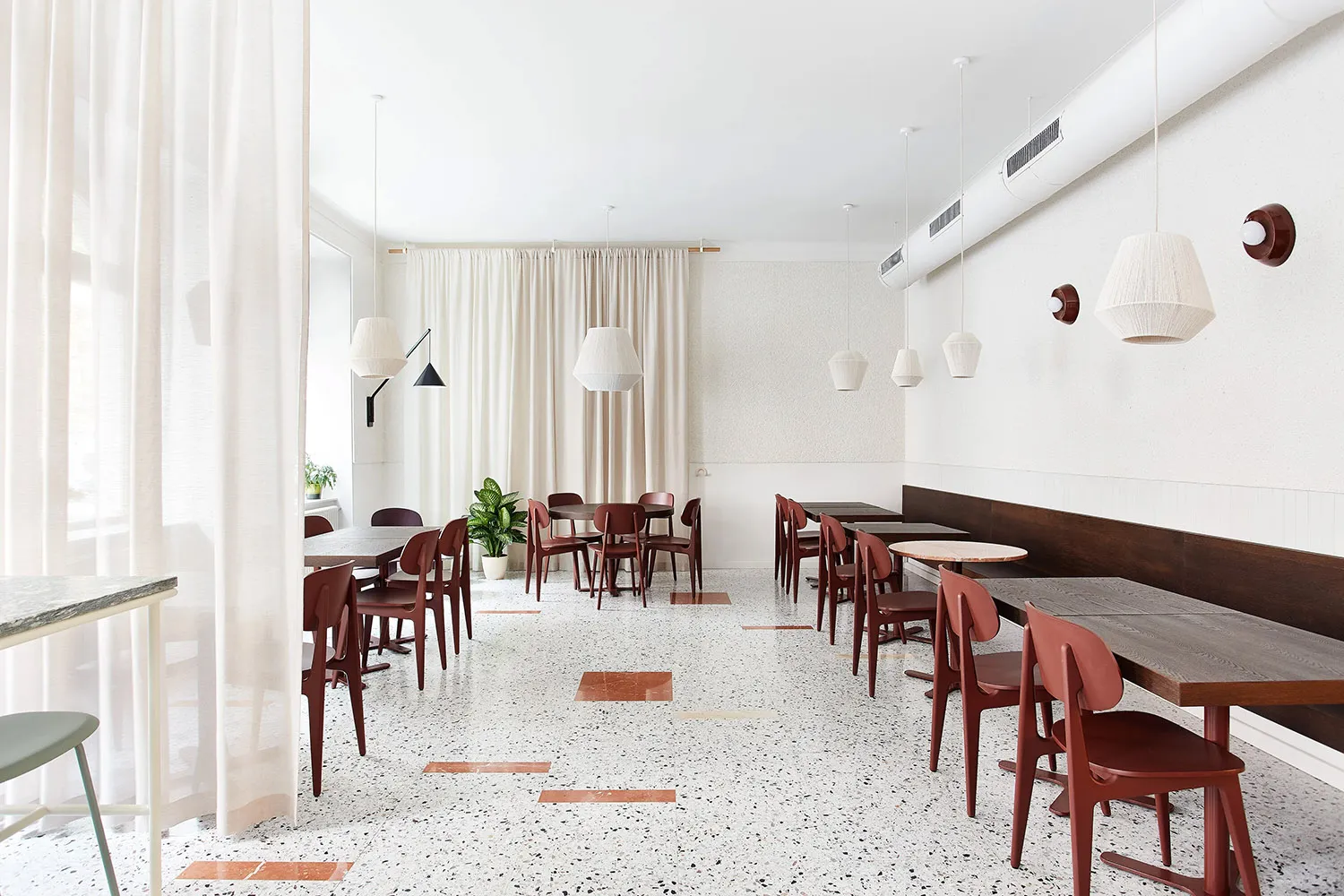 A minimalist restaurant interior with terrazzo flooring, wooden tables, and red chairs.