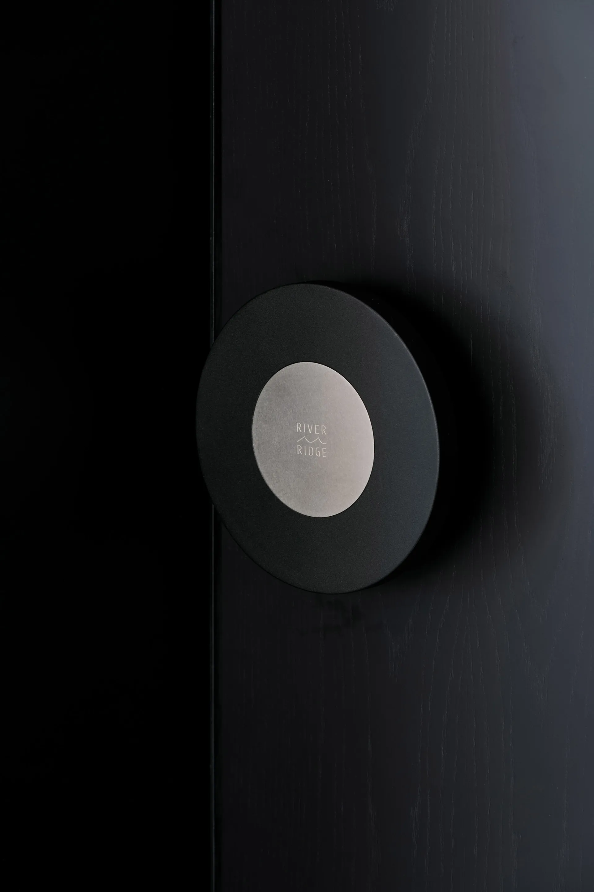 A minimalist, black, round, metallic door handle against a black, textured wall background.