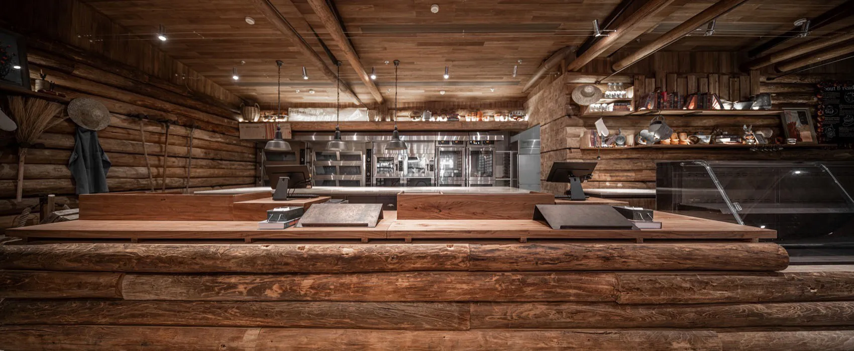 A spacious, rustic restaurant interior with exposed wooden logs on the walls and ceiling, and a modern kitchen area in the ba